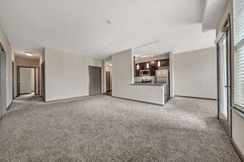 A spacious, empty room with a carpeted floor and a kitchen area in the background.